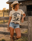 Woman wearing sand colored A Hard Head Makes a Soft Ass western graphic t-shirt styled with denim shorts, cowboy boots, and cowboy hat outdoors