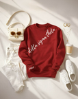 Flat lay photo of a crimson red sweatshirt with white diagonal script reading “delta sigma theta,” styled with white ripped jeans, white sneakers, gold jewelry, sunglasses, a cream handbag, and a coffee cup on a light neutral background, creating a chic casual sorority fashion aesthetic.