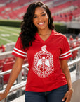A smiling woman stands in a stadium wearing a bright red V-neck athletic jersey with white sleeve stripes and a large white Greek letter crest on the front, paired with dark blue jeans.
