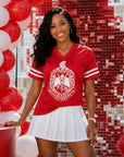 A woman stands in a festive red and white decorated setting, wearing a red short-sleeve athletic-style T-shirt with white sleeve stripes and a large white crest graphic on the front, paired with a white pleated mini skirt.