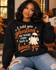 A woman wearing a black crewneck sweatshirt with a Halloween graphic that reads 'I told you ghosting me would come back to haunt you' in orange and white, paired with ripped blue jeans, sitting in a cozy living room setting.