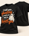 A black short-sleeve graphic T-shirt shown from the front and back on a light beige background. The front features a Halloween-themed print with the phrase 'I told you ghosting me would come back to haunt you' in orange and white lettering, alongside a white ghost illustration and small white stars.