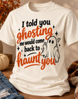 An off-white short-sleeve graphic T-shirt displayed in a styled autumn setting on a wooden surface, with a pumpkin, denim, and a plaid blanket nearby. The shirt features the text 'I told you ghosting me would come back to haunt you' in black and orange lettering, alongside a simple black-and-white ghost illustration.