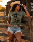 Black woman wearing olive green A Hard Head Makes a Soft Ass western graphic t-shirt with denim shorts, cowboy boots, and cowboy hat at sunset ranch setting