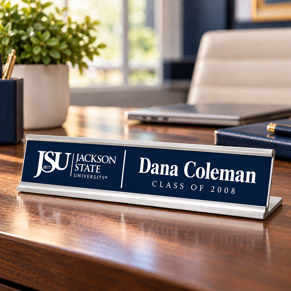 A personalized nameplate sign on a wooden desk, featuring a navy blue front with white text that reads 'JSU Jackson State University' and 'Dana Coleman Class of 2008'. The sign has a polished silver metal base and is shown in a blurred office environment with a plant, laptop, and chair in the background.