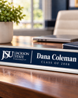 A personalized nameplate sign on a wooden desk, featuring a navy blue front with white text that reads 'JSU Jackson State University' and 'Dana Coleman Class of 2008'. The sign has a polished silver metal base and is shown in a blurred office environment with a plant, laptop, and chair in the background.