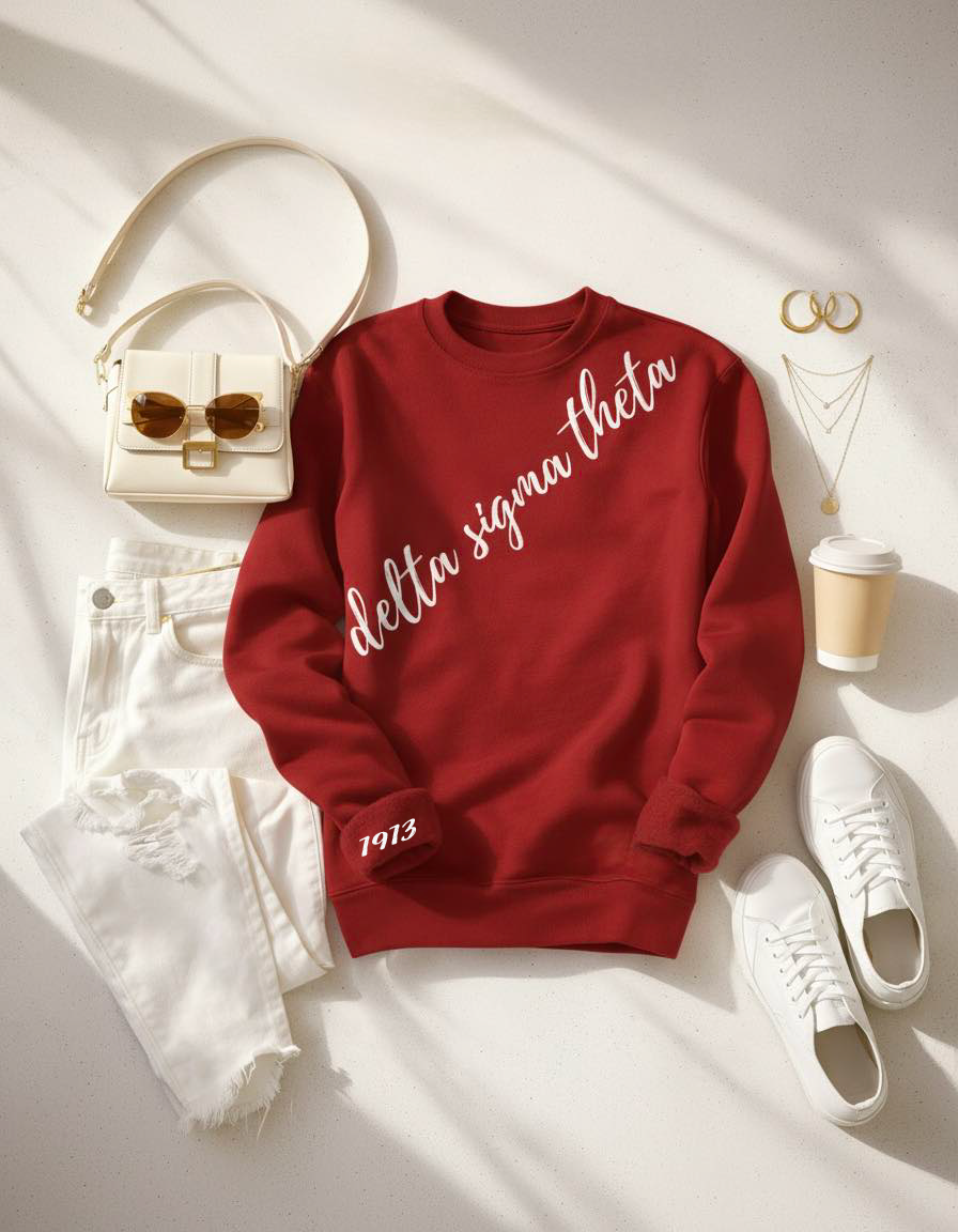 Flat lay photo of a crimson red sweatshirt with white diagonal script reading “delta sigma theta,” styled with white ripped jeans, white sneakers, gold jewelry, sunglasses, a cream handbag, and a coffee cup on a light neutral background, creating a chic casual sorority fashion aesthetic.
