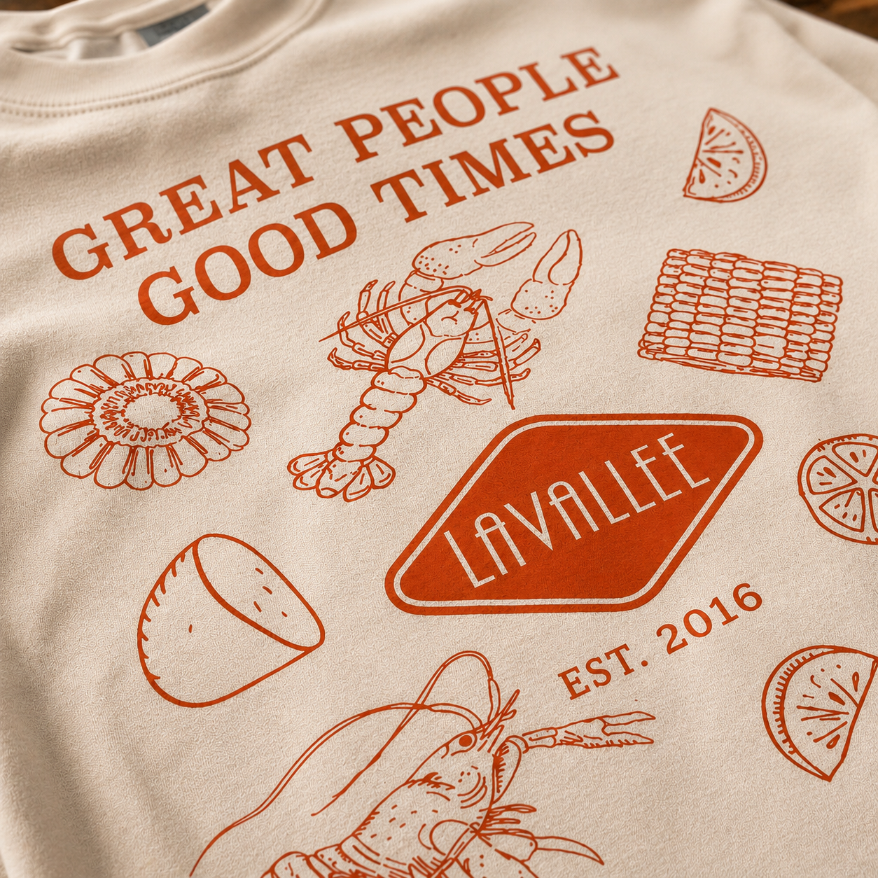 Close-up of Lavallee anniversary shirt graphic featuring crawfish, corn, citrus illustrations, and “Great People Good Times” typography in burnt orange ink on cream t-shirt.