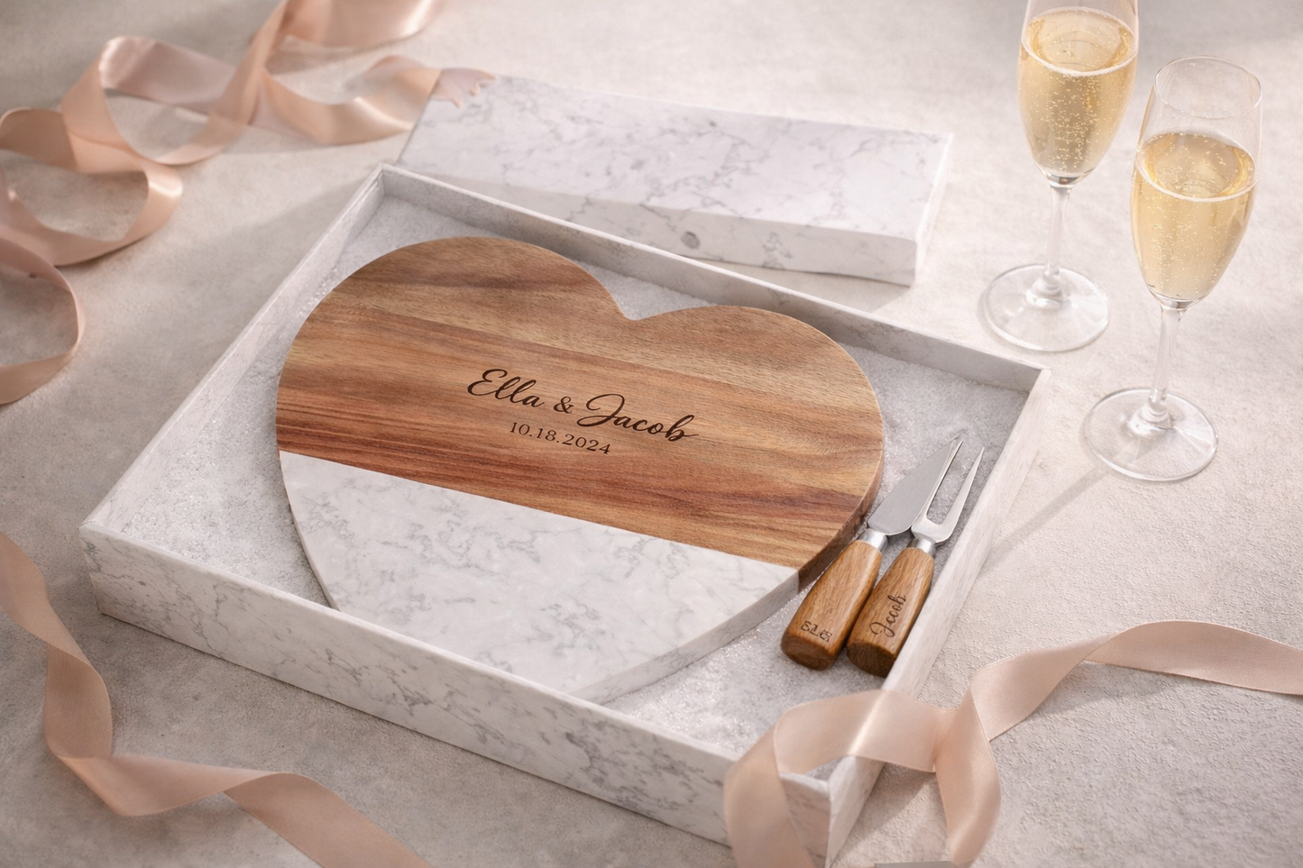 Heart-shaped cutting board with engraved names and date, accompanied by a knife and fork set, on a light gray surface with ribbons and champagne glasses.