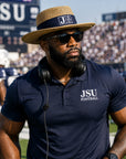 A man on a football field wearing a navy blue JSU Football polo shirt, a straw block letter JSU gambler hat, and sunglasses, with players and stadium stands blurred in the background.