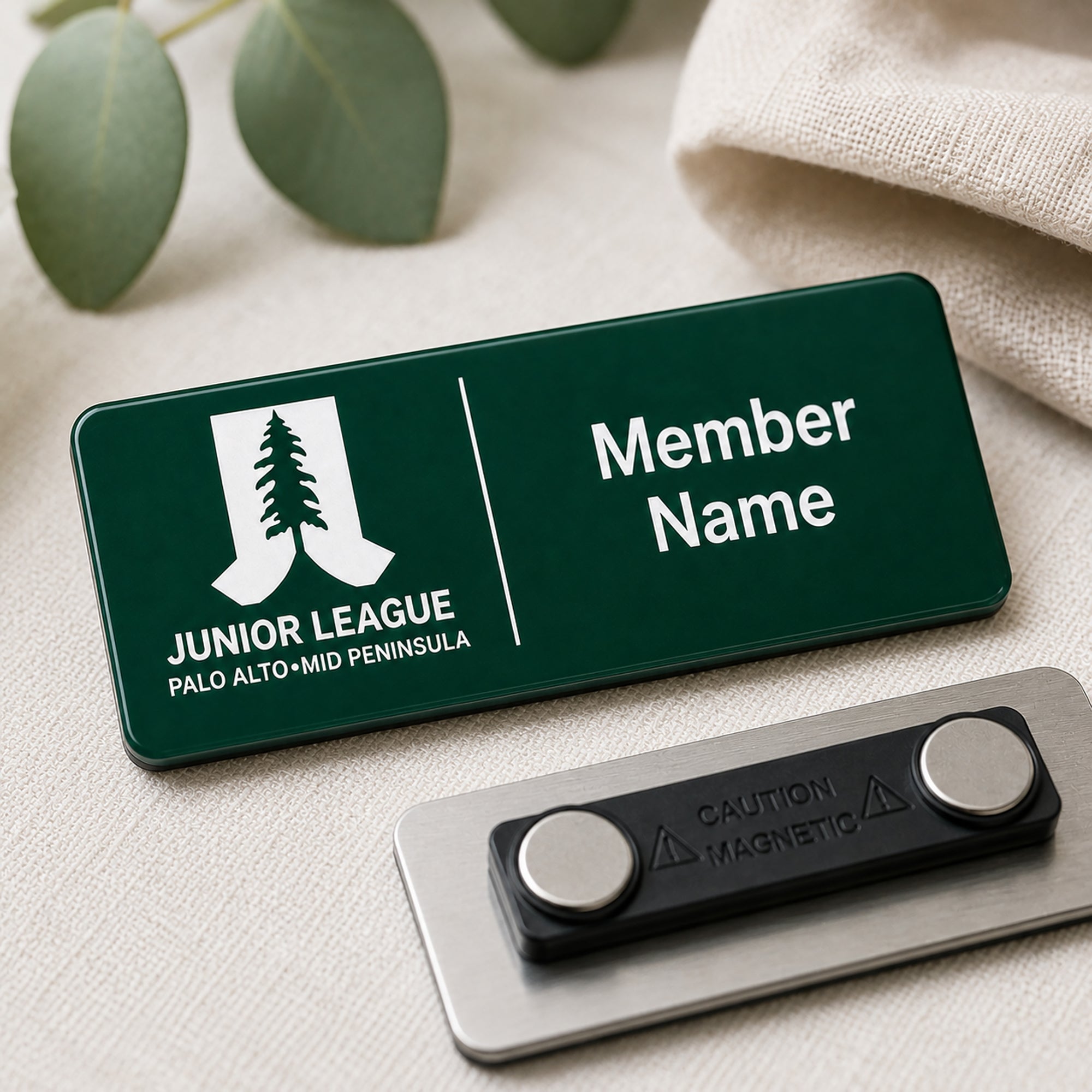 Laser engraved green and white two-tone acrylic Junior League Palo Alto Mid Peninsula custom member name badge with magnetic back shown on linen surface with eucalyptus leaves