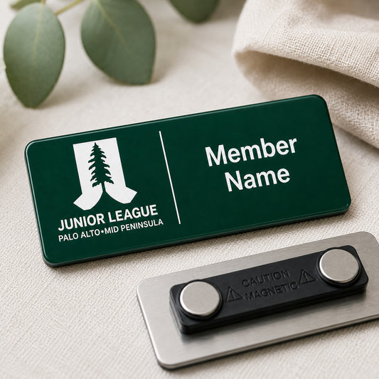 Laser engraved green and white two-tone acrylic Junior League Palo Alto Mid Peninsula custom member name badge with magnetic back shown on linen surface with eucalyptus leaves