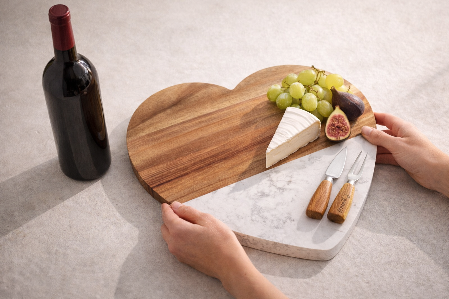 Heart-shaped cutting board with cheese, grapes, figs, and a bottle of wine on a light surface.
