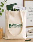 Natural tote bag with Junior League logo on a wooden surface with books and a drink.