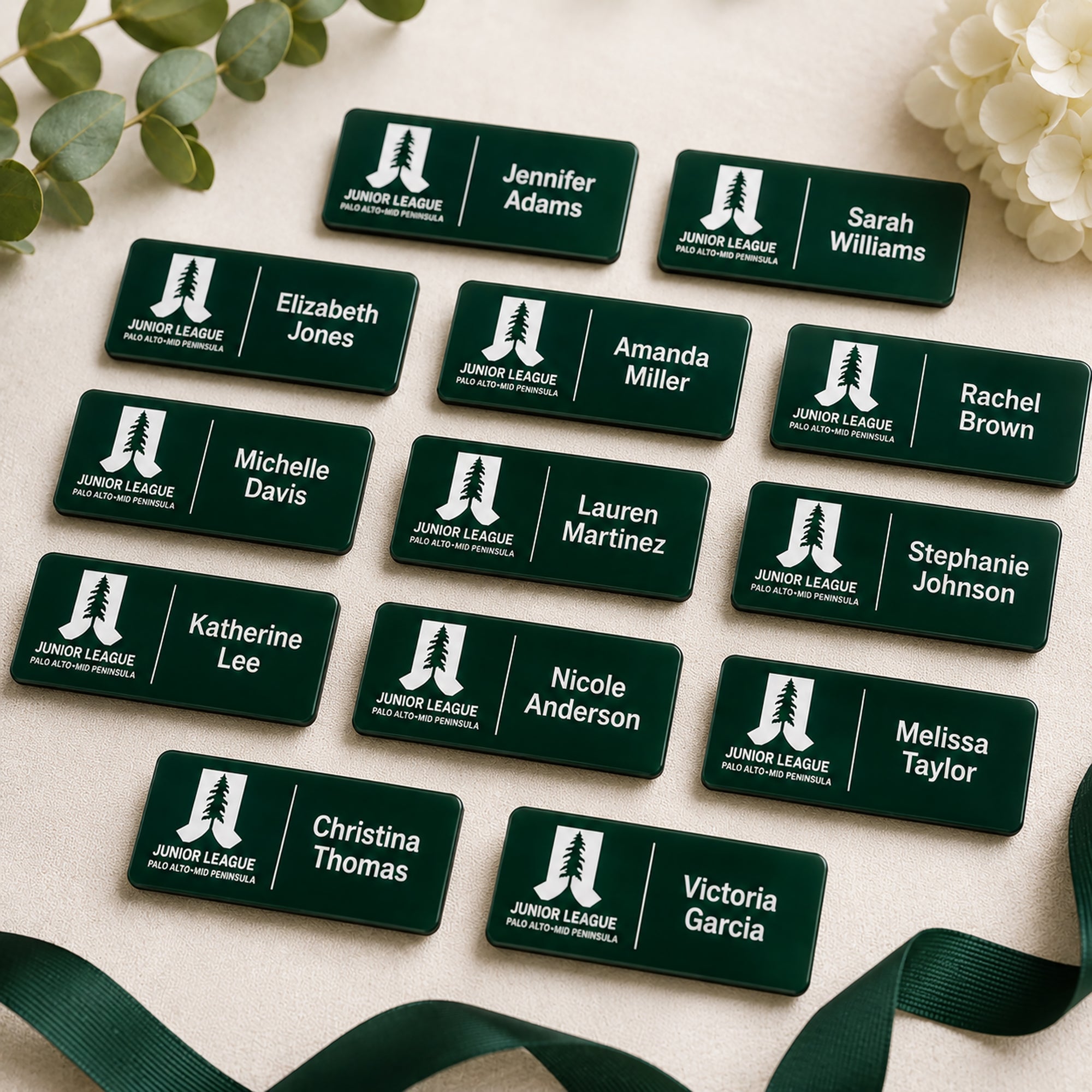 Flat lay of multiple laser engraved Junior League Palo Alto Mid Peninsula custom name badges personalized with member names arranged on neutral linen background with eucalyptus and white flowers