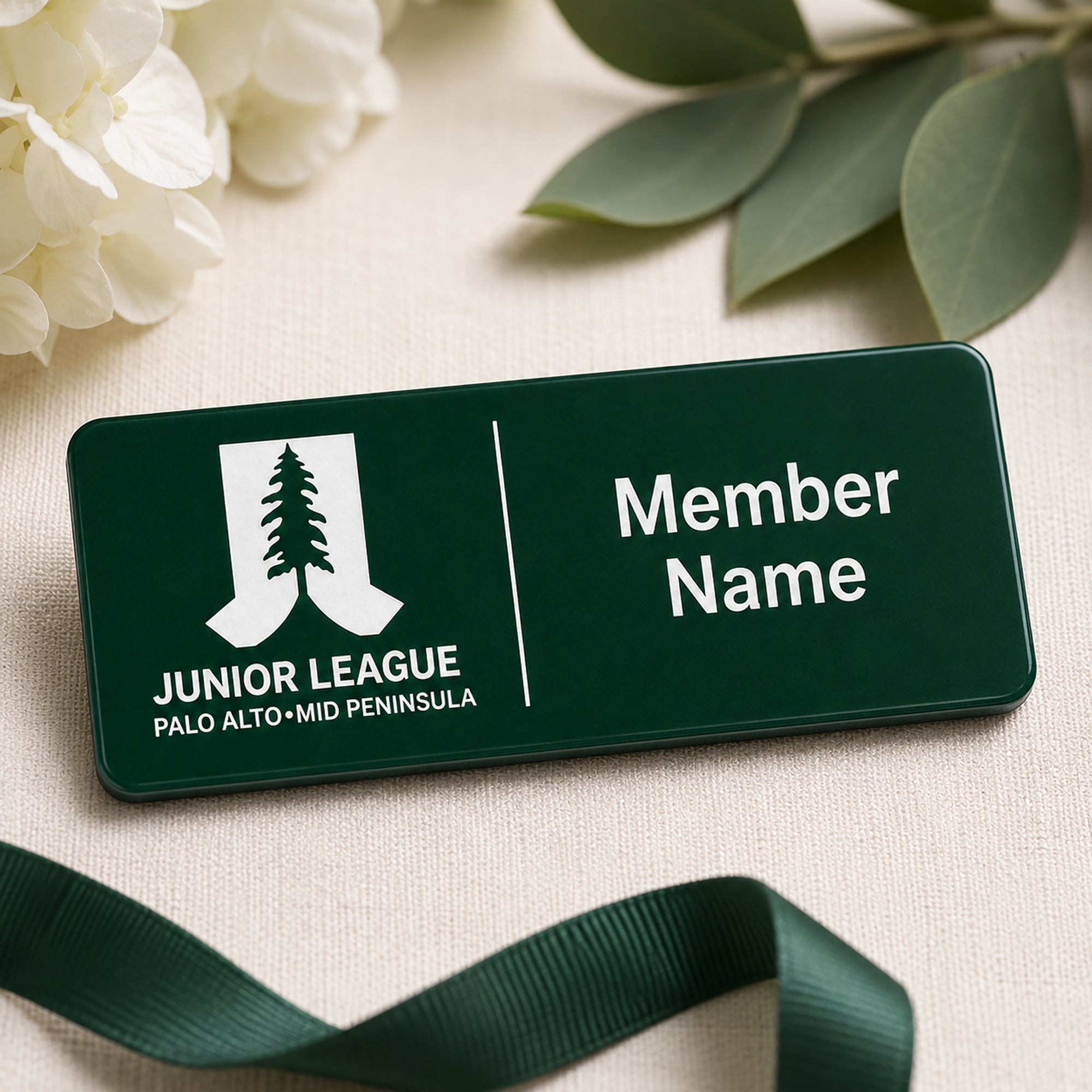 Close-up of laser engraved Junior League Palo Alto Mid Peninsula green acrylic name badge with white engraving and dark green grosgrain ribbon on linen background with white flowers