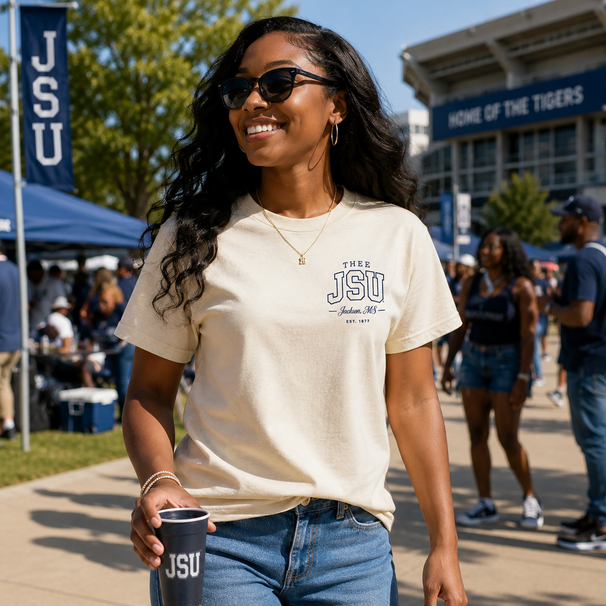 Woman wearing a beige t-shirt with 'JSU' on it, holding a cup, and standing in a casual outdoor setting.