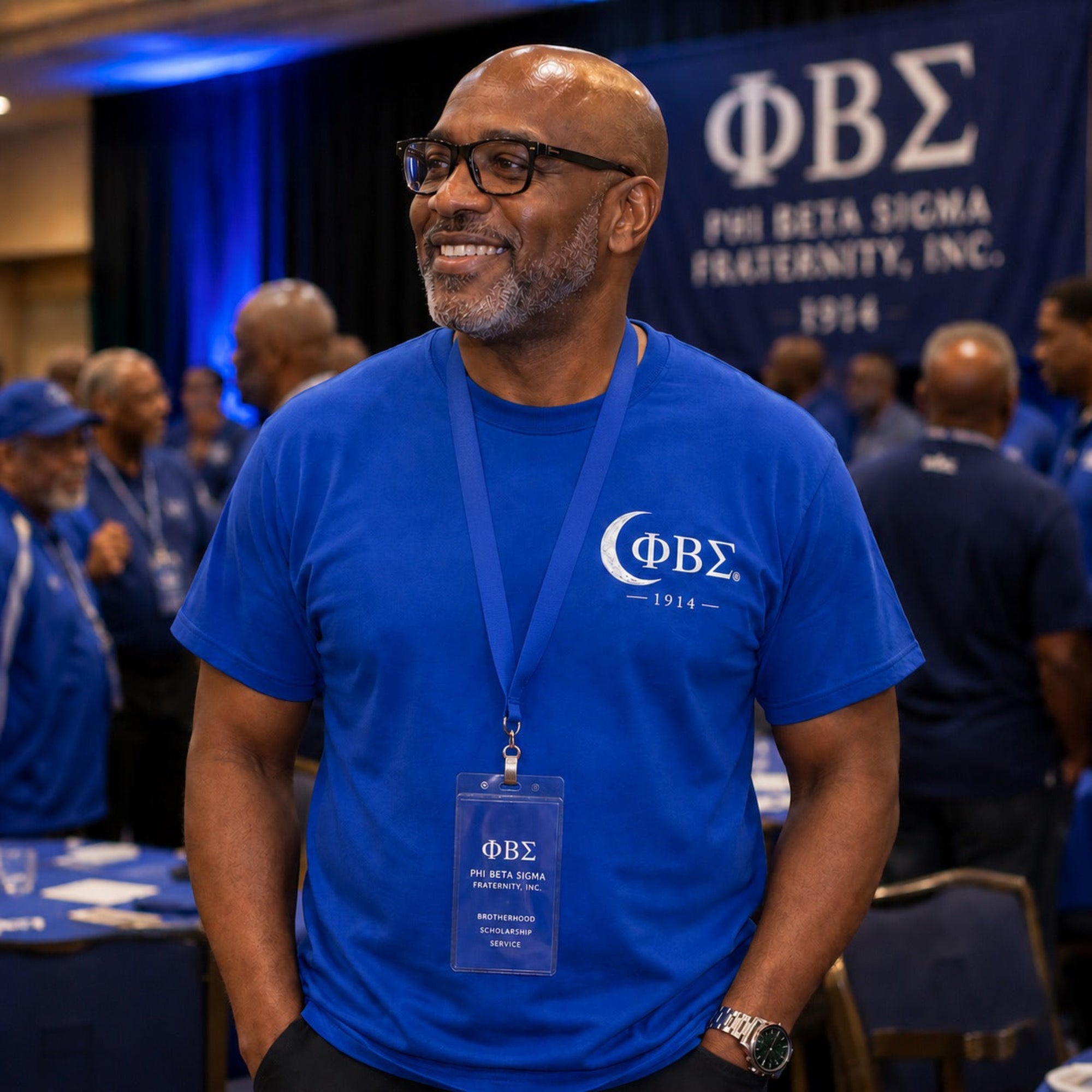 Man wearing a blue Phi Beta Sigma t-shirt with a lanyard, standing in front of a Phi Beta Sigma banner.
