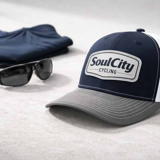 Navy and white SoulCity Cycling trucker hat on concrete surface with sunglasses and folded shirt