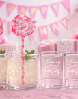 Clear shot glasses with 'Blooming in my Forties' text, pink straws, and decorative background with balloons and gift boxes.