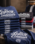 Stacks of t-shirts with 'The Legacy Continues' text on a wooden table.