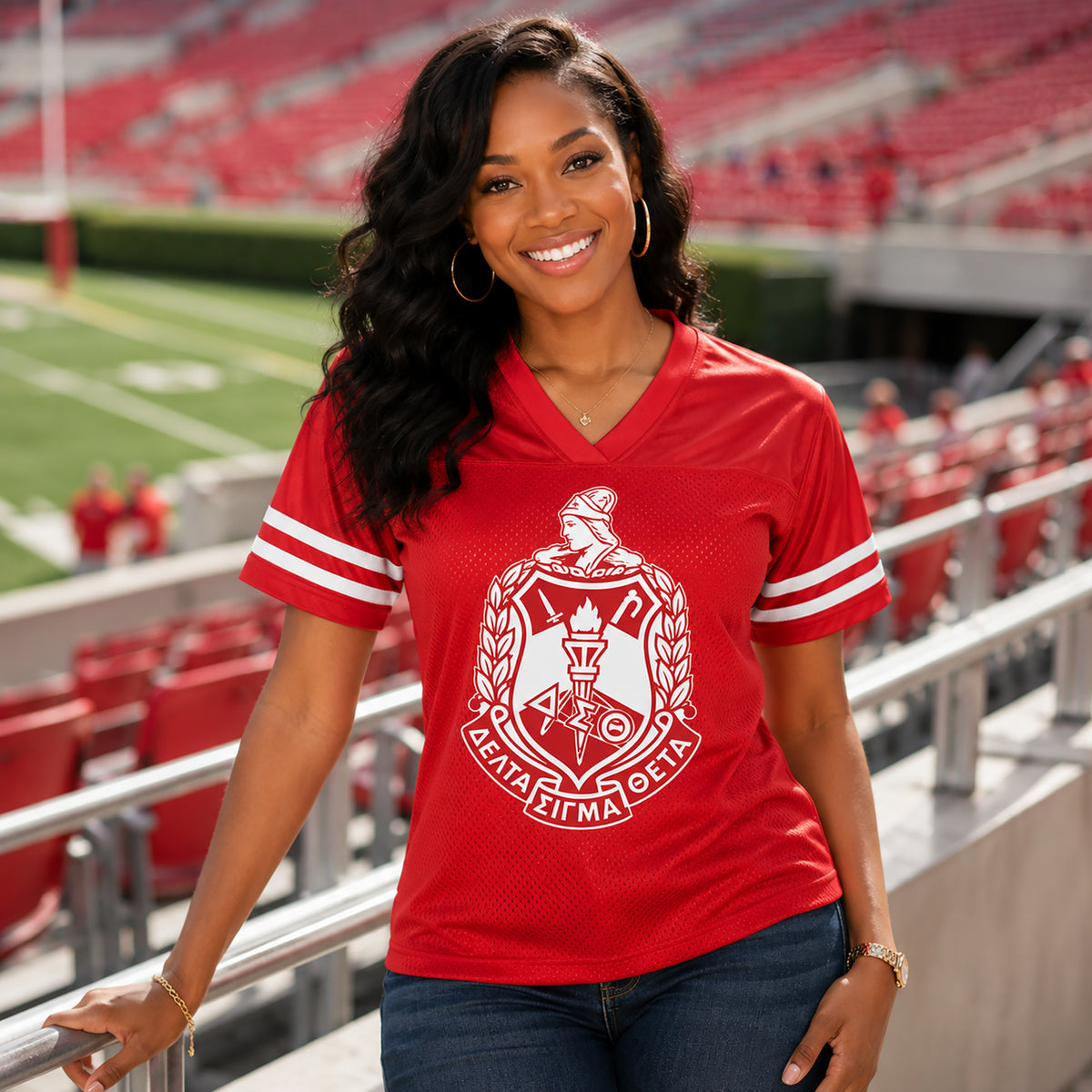 A smiling woman stands in a stadium wearing a bright red V-neck athletic jersey with white sleeve stripes and a large white Greek letter crest on the front, paired with dark blue jeans.