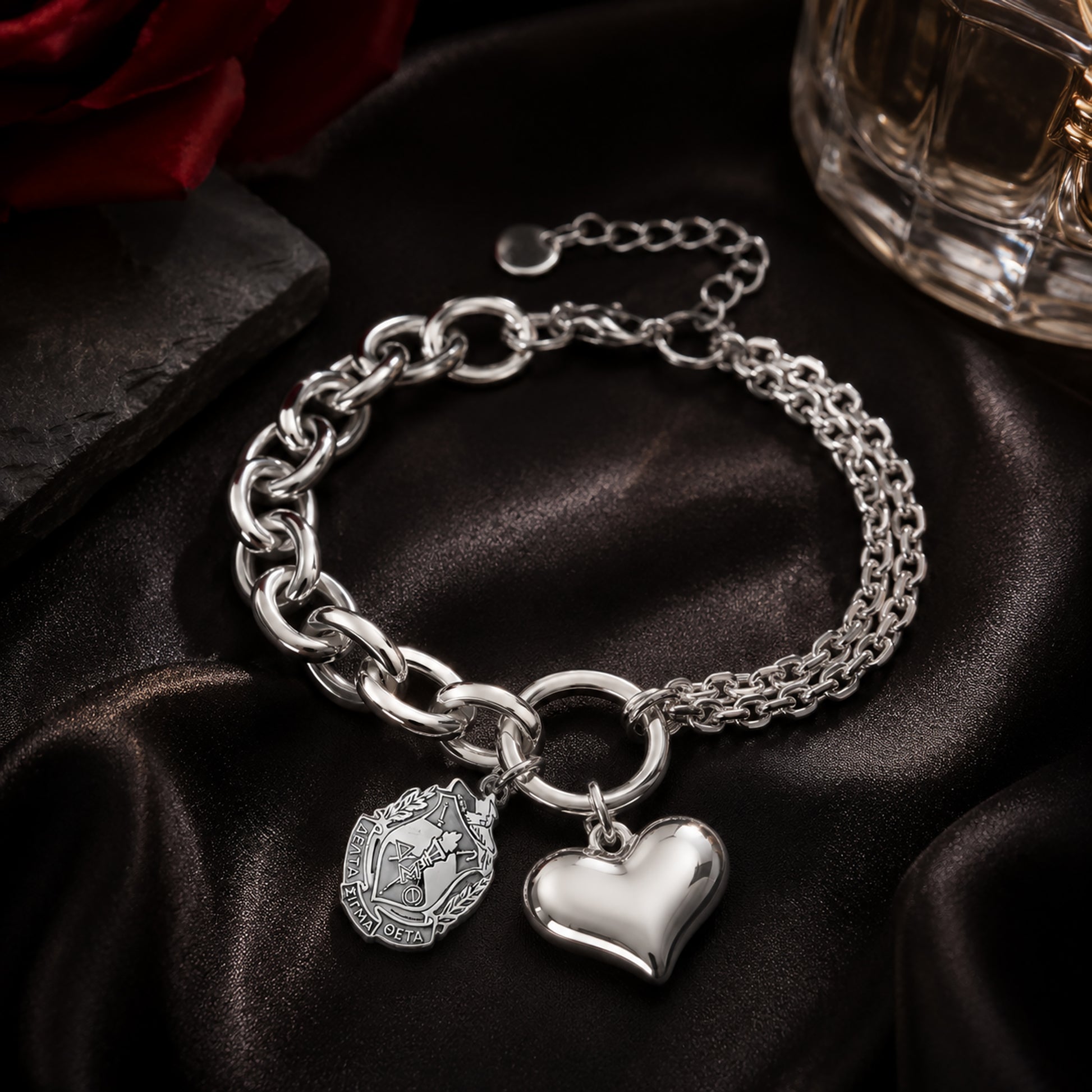 Officially licensed Delta Sigma Theta shield charm bracelet featuring a polished stainless steel heart charm displayed on black satin with a crimson rose and luxury perfume bottle.