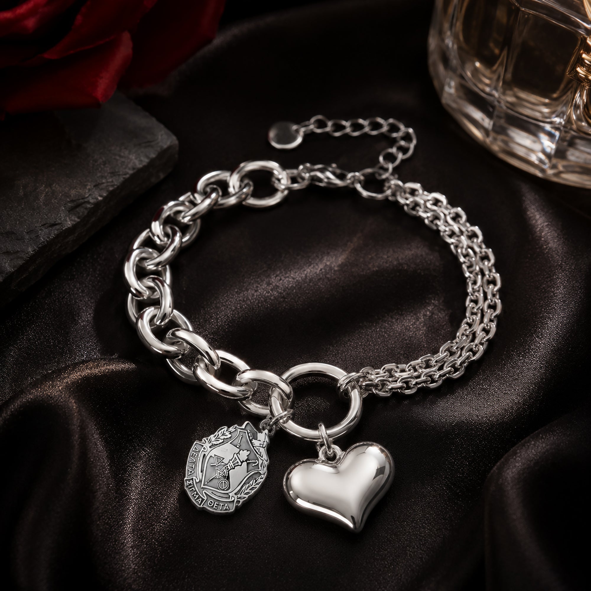 Officially licensed Delta Sigma Theta shield charm bracelet featuring a polished stainless steel heart charm displayed on black satin with a crimson rose and luxury perfume bottle.
