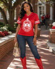 A woman stands outdoors on a brick walkway wearing a bright red V-neck graphic T-shirt with white sleeve stripes and a large crest print, paired with dark blue jeans, red cowboy boots, and holding a clear handbag.