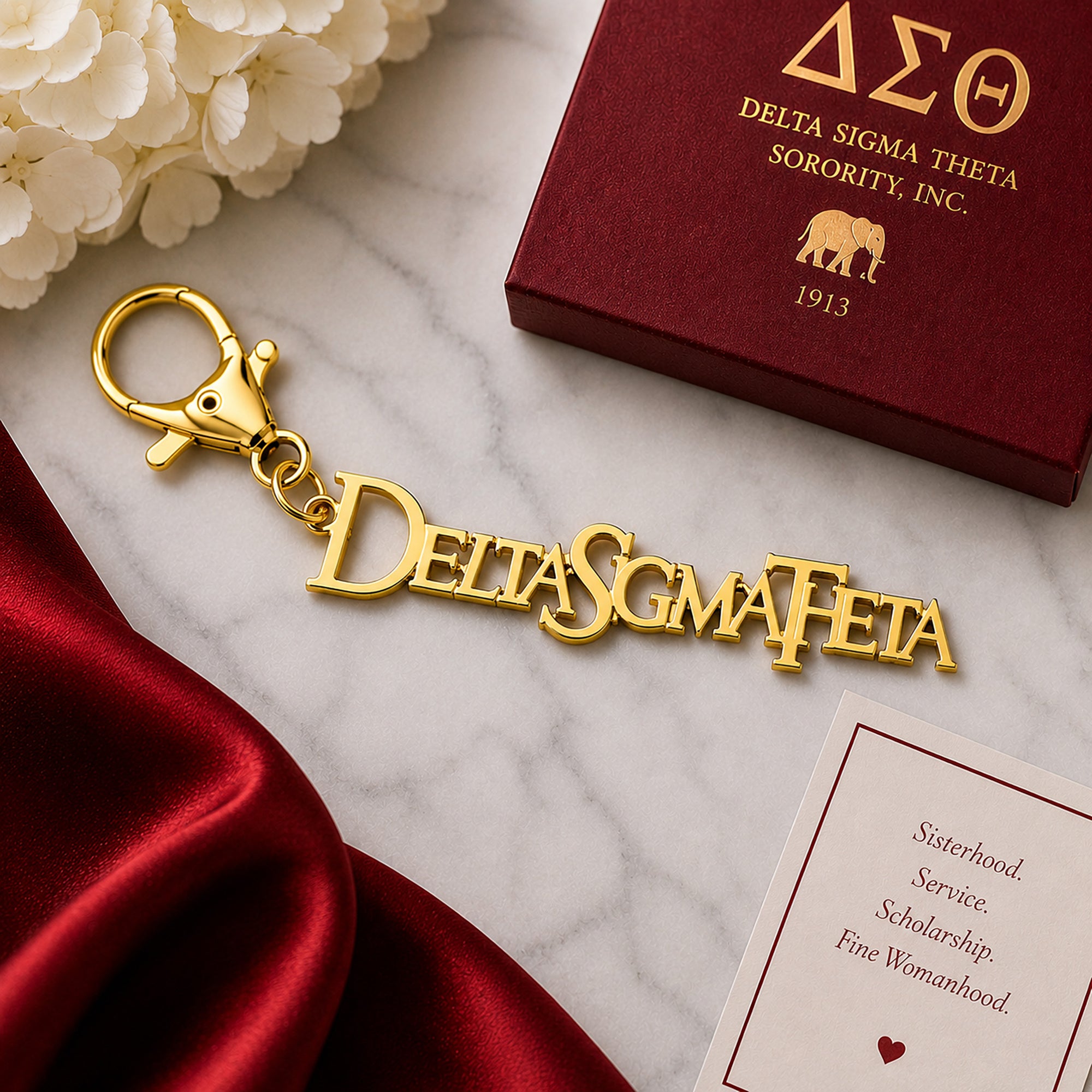 Officially licensed Delta Sigma Theta script logo keychain in a polished gold finish with lobster clasp displayed on a marble surface with luxury gift packaging and elegant red satin accents.