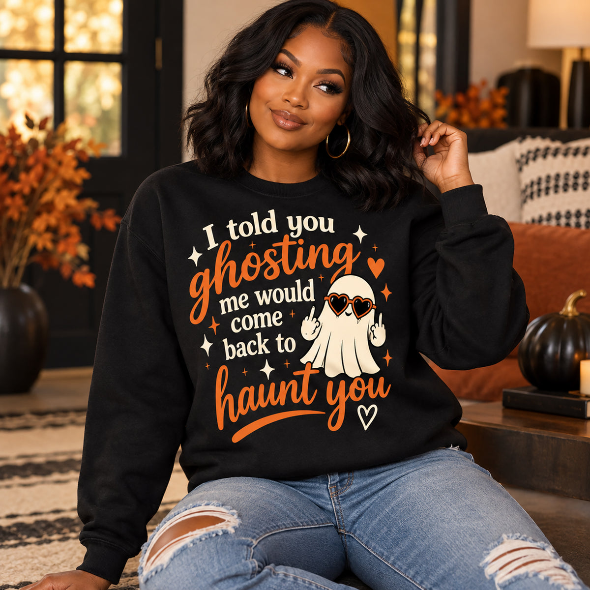 A woman wearing a black crewneck sweatshirt with a Halloween graphic that reads 'I told you ghosting me would come back to haunt you' in orange and white, paired with ripped blue jeans, sitting in a cozy living room setting.