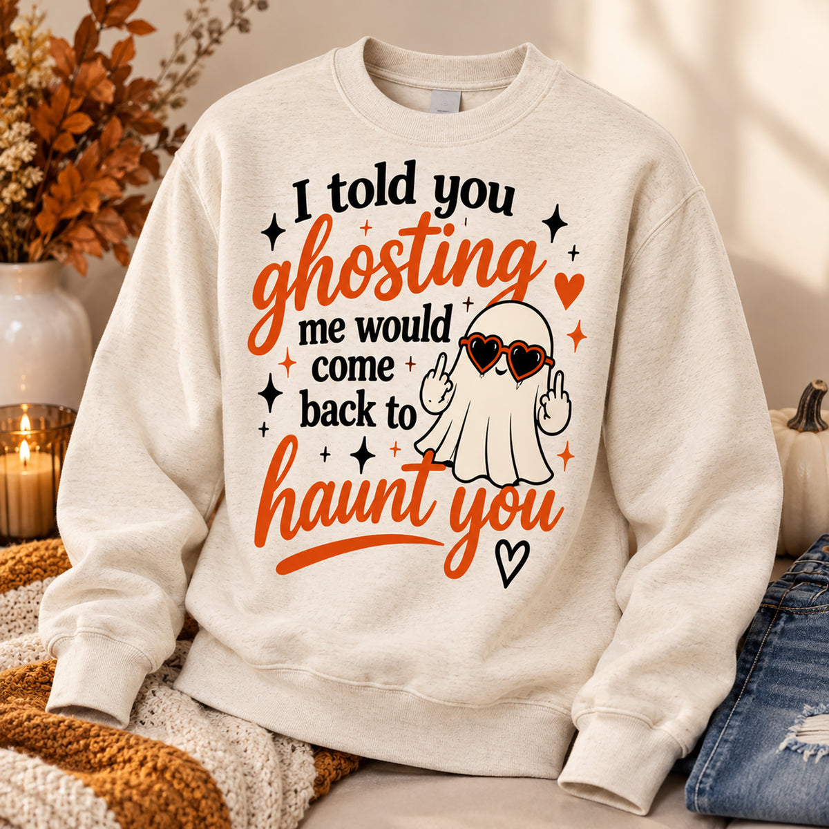An off-white crewneck sweatshirt with a Halloween graphic print, styled in a cozy autumn setting with a knit blanket, a candle, and denim jeans.