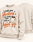 A light beige crewneck sweatshirt shown from both front and back. The front features a Halloween-themed graphic with the text 'I told you ghosting me would come back to haunt you' in black and orange lettering, a cartoon ghost wearing heart-shaped sunglasses, and small orange and black stars.