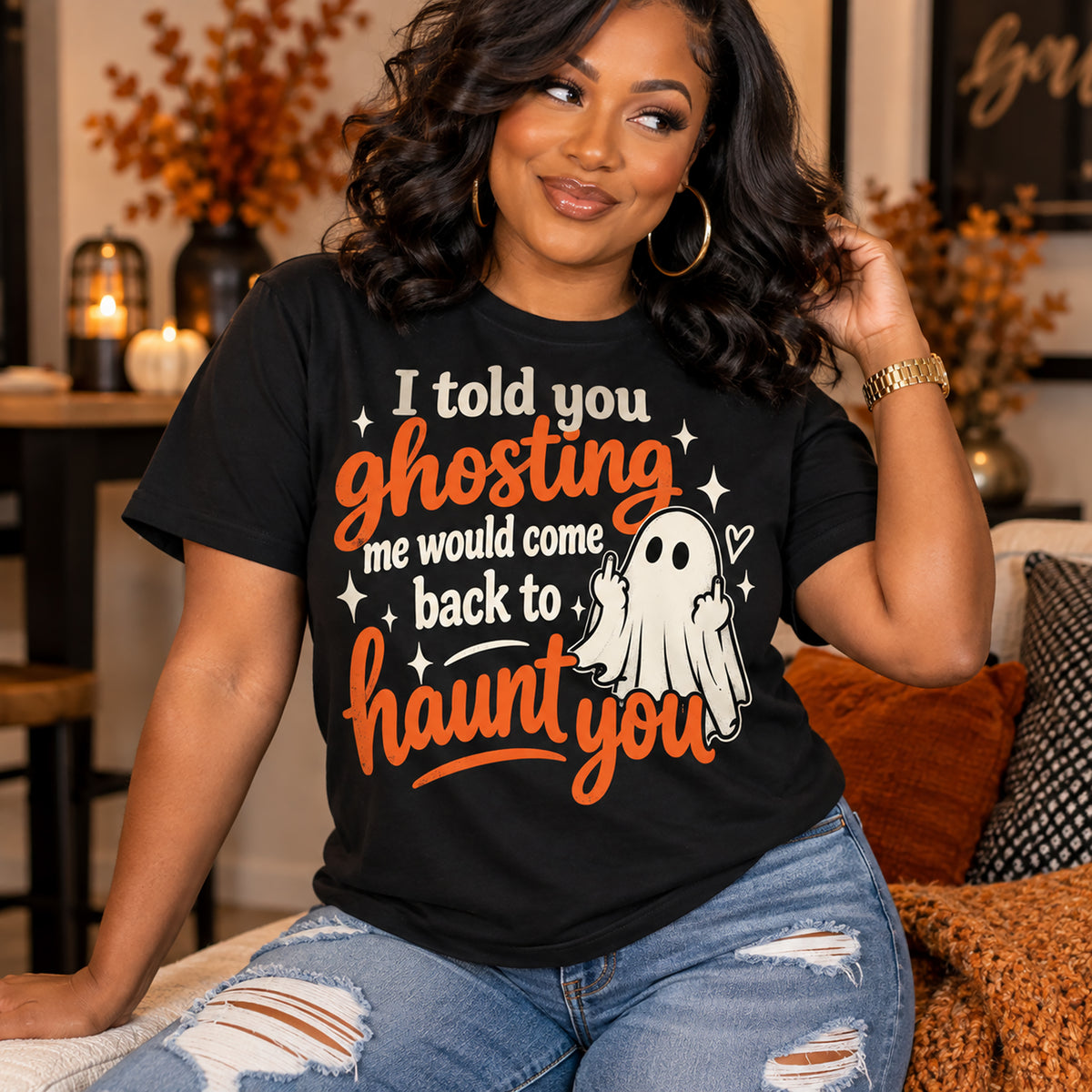 A woman wearing a black graphic t-shirt with a Halloween-themed design that reads 'I told you ghosting me would come back to haunt you' alongside a cartoon ghost illustration. She is sitting on a couch in a cozy, warmly lit living room.