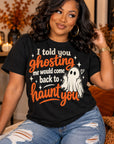 A woman wearing a black graphic t-shirt with a Halloween-themed design that reads 'I told you ghosting me would come back to haunt you' alongside a cartoon ghost illustration. She is sitting on a couch in a cozy, warmly lit living room.