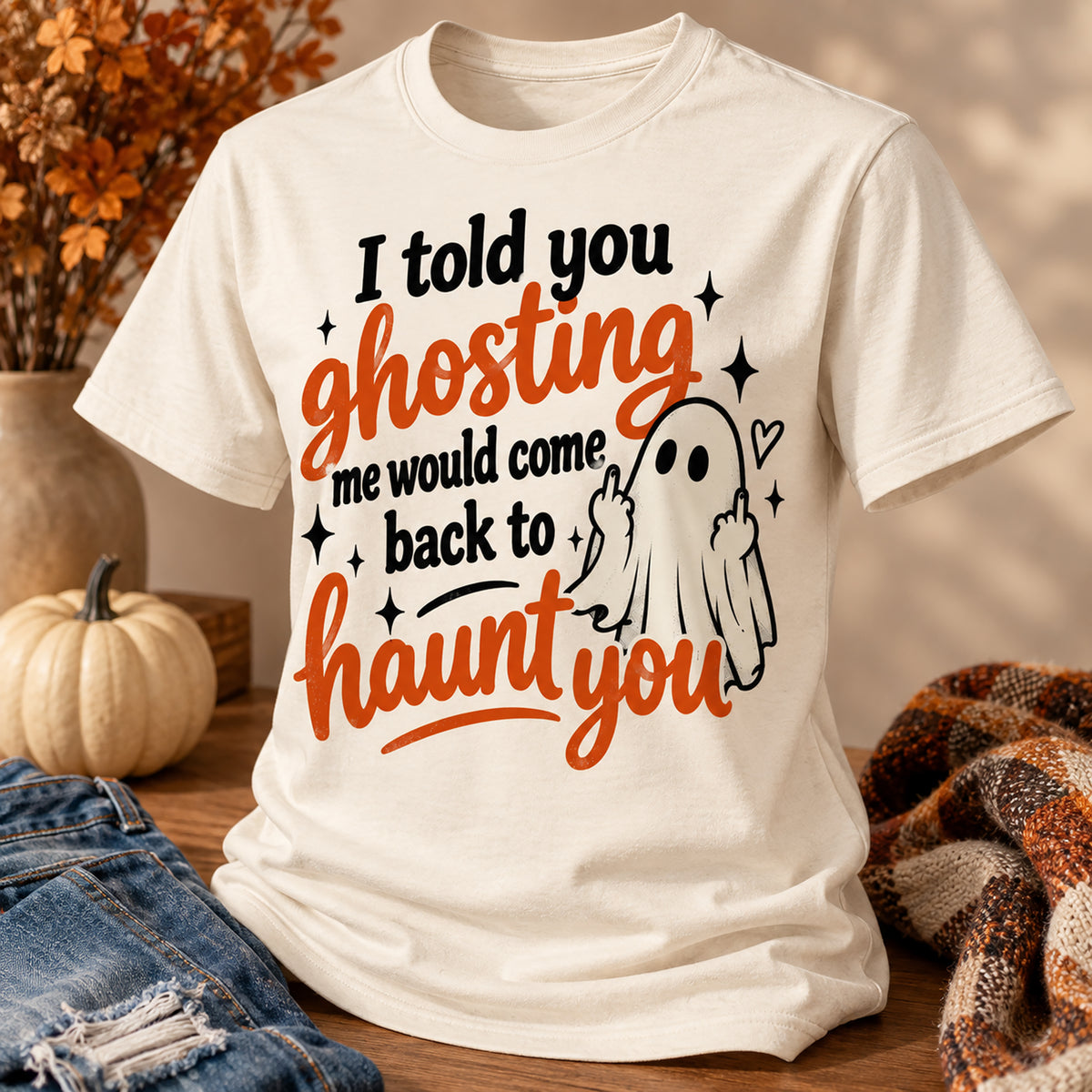 An off-white short-sleeve graphic T-shirt displayed in a styled autumn setting on a wooden surface, with a pumpkin, denim, and a plaid blanket nearby. The shirt features the text 'I told you ghosting me would come back to haunt you' in black and orange lettering, alongside a simple black-and-white ghost illustration.