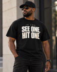 A man wearing a black t-shirt with the bold white text 'IF YOU SEE ONE, HIT ONE.' printed on the chest, styled with a black baseball cap, sunglasses, and dark jeans in an outdoor urban setting.