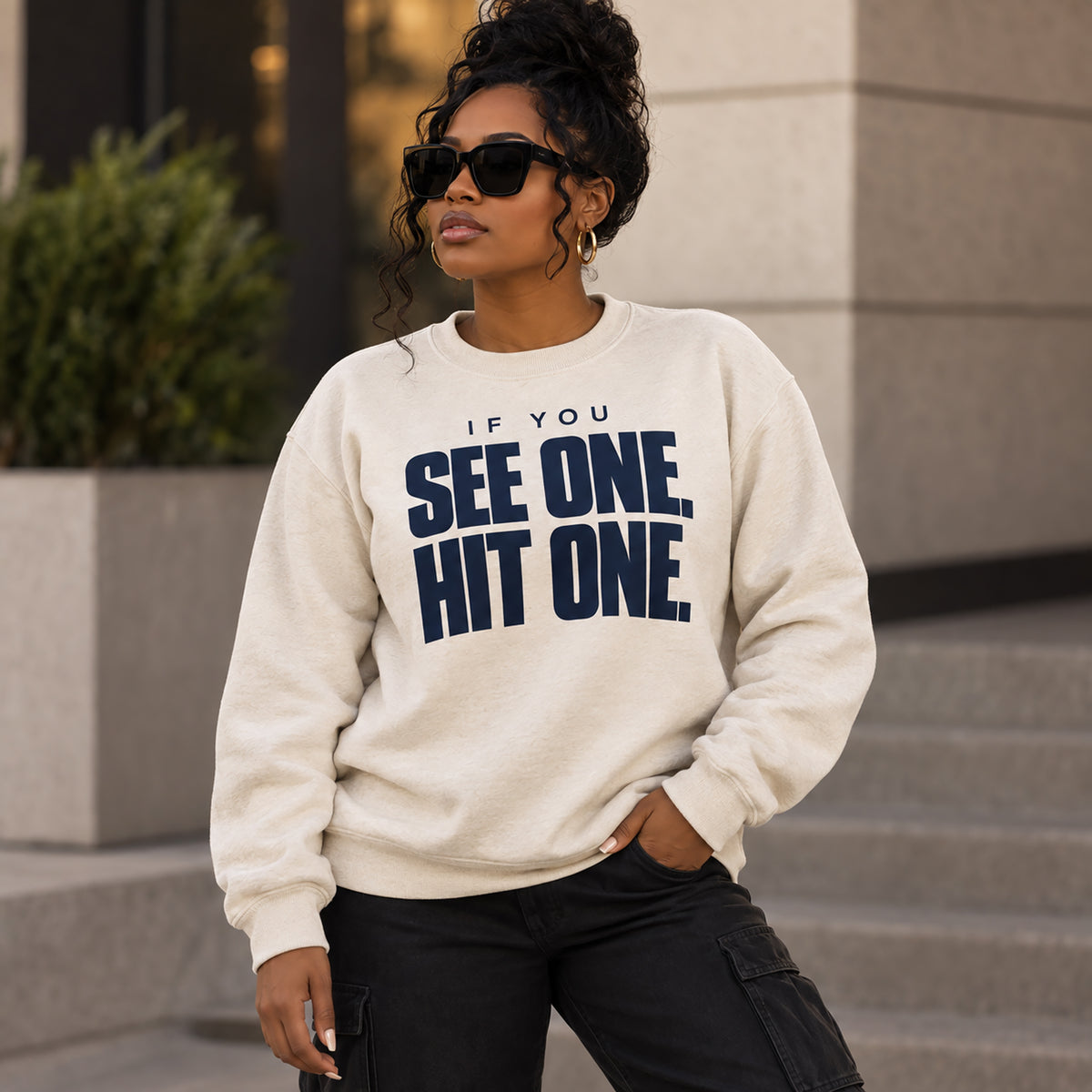 A woman wearing a cream-colored crewneck sweatshirt with the text 'IF YOU SEE ONE, HIT ONE.' printed in bold navy blue letters, paired with black cargo pants and sunglasses, standing outdoors in front of a modern building.