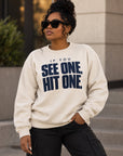 A woman wearing a cream-colored crewneck sweatshirt with the text 'IF YOU SEE ONE, HIT ONE.' printed in bold navy blue letters, paired with black cargo pants and sunglasses, standing outdoors in front of a modern building.