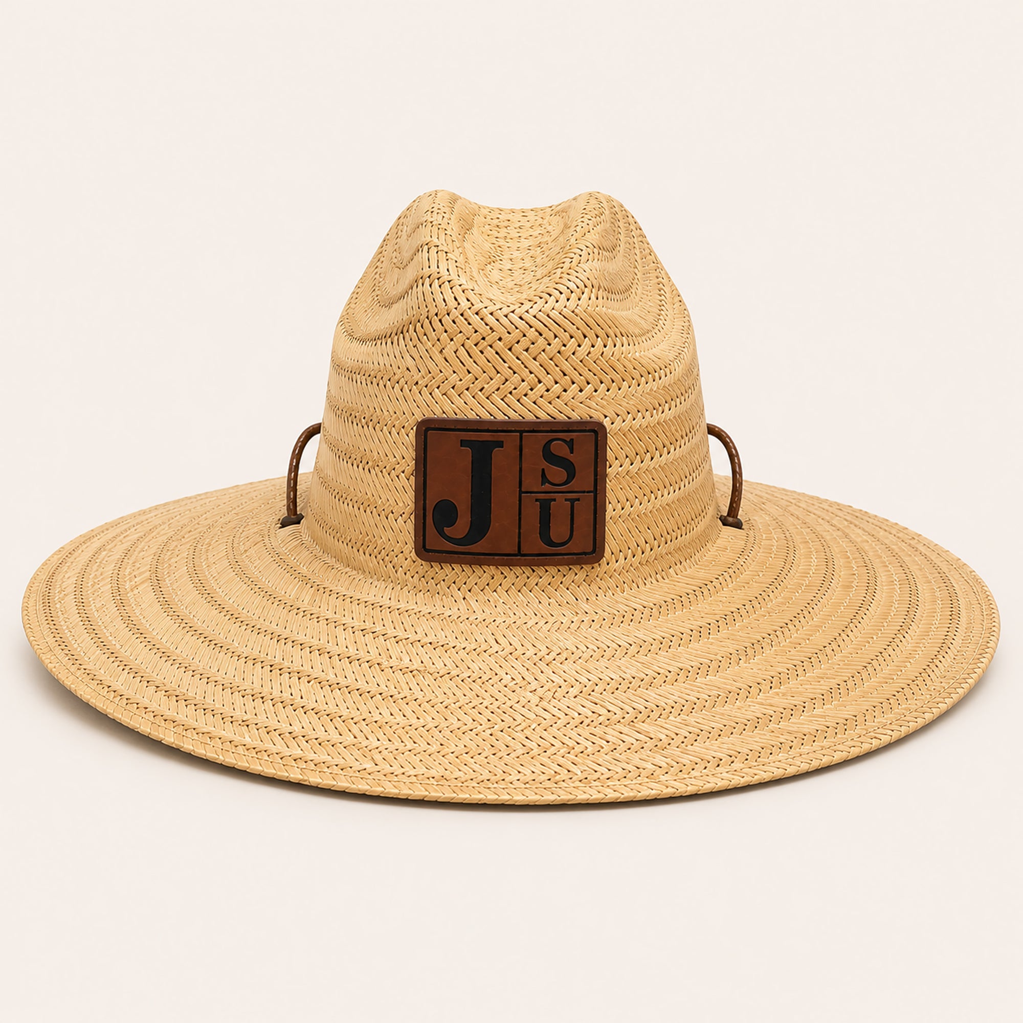A woven straw safari-style hat in a light tan color, featuring a brown leather patch on the front with the letters 'J', 'S', and 'U' in black, and two small brown side loops.
