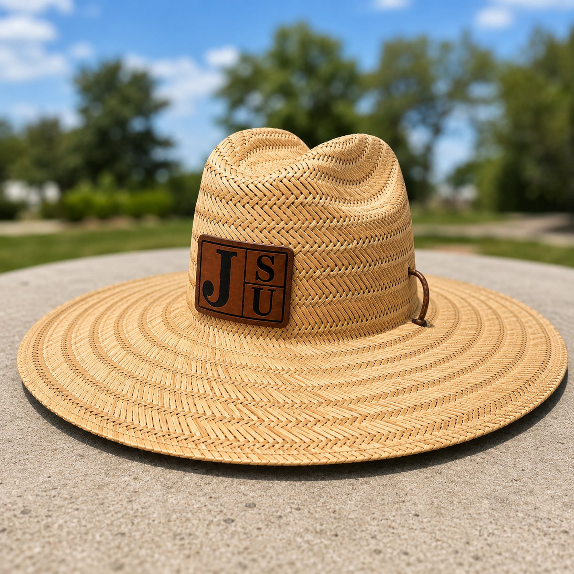 A natural straw wide-brim hat with a brown leather patch featuring the letters 'J S U' on a sunny outdoor day, set against a blurred background of green trees and blue sky.