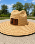 A natural straw wide-brim hat with a brown leather patch featuring the letters 'J S U' on a sunny outdoor day, set against a blurred background of green trees and blue sky.