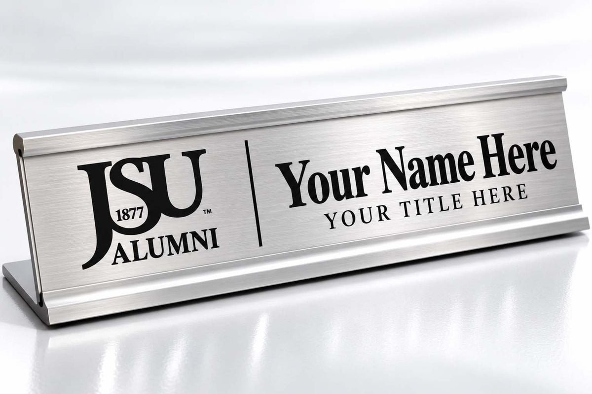 A brushed stainless steel tabletop nameplate with black engraved text reading 'JSU ALUMNI' and 'Your Name Here / YOUR TITLE HERE', shown on a clean white reflective surface.