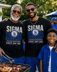 Grandfather, dad, and son wearing 'Sigma Dad' shirts standing around a grill outdoors.