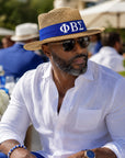 A man wearing a straw fedora hat with a royal blue band and Greek letters ΦΒΣ, a white linen shirt, and sunglasses, seated outdoors at a social event.